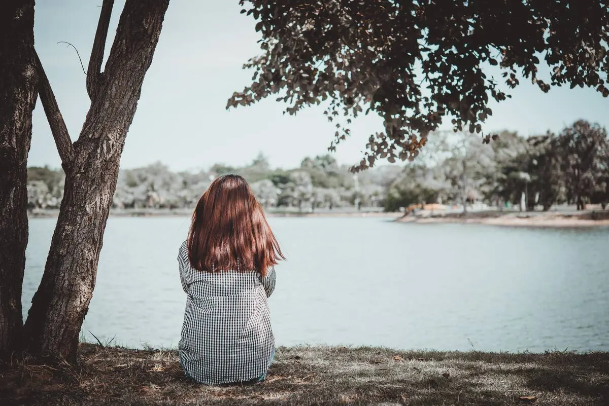 A woman sitting alone under a tree, looking out over a quiet lake
