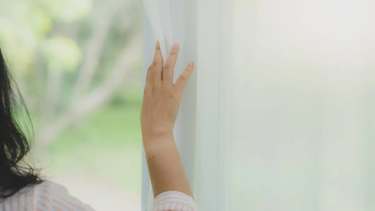 A hand drawing back a sheer white curtain, soft green daylight beyond